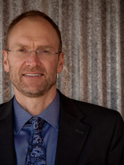 photo of a a man from the shoulders up wearing a blue shirt, tie, and black jacket