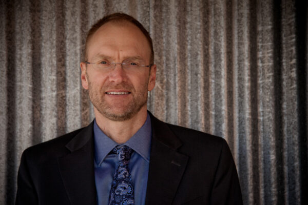 photo of a a man from the shoulders up wearing a blue shirt, tie, and black jacket