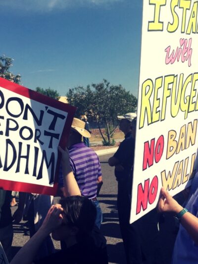 Protesters hold posters that read "Don't deport Kadhim" and "I stand with refugees.  No ban. No wall."