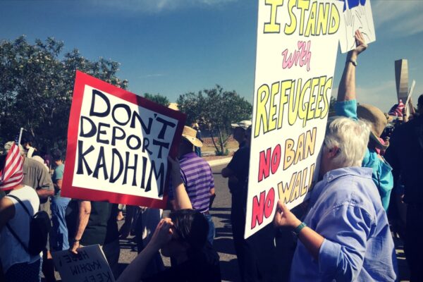 Protesters hold posters that read "Don't deport Kadhim" and "I stand with refugees.  No ban. No wall."