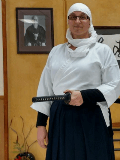A woman stands wearing a white headscarf and martial arts robe, holding a sword
