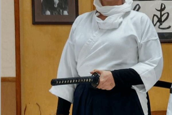 A woman stands wearing a white headscarf and martial arts robe, holding a sword