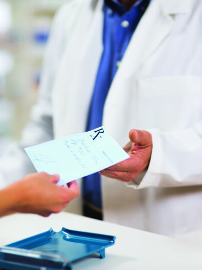 Photo: cropped photo of a pharmacist in a white coat behind a counter receiving a paper prescription from an outstretched hand