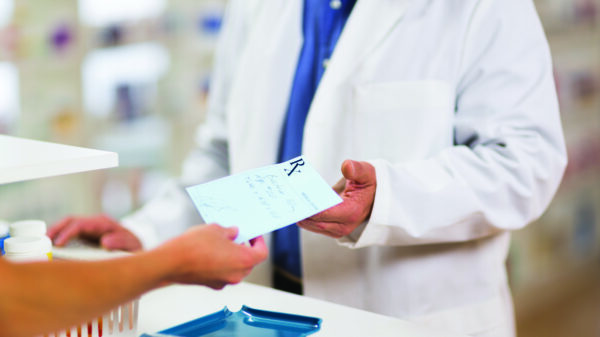 Photo: cropped photo of a pharmacist in a white coat behind a counter receiving a paper prescription from an outstretched hand