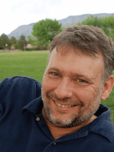 Photo: A man with short hair and groomed facial hair in a navy blue polo shirt reclining on a field of grass, smiling at the camera