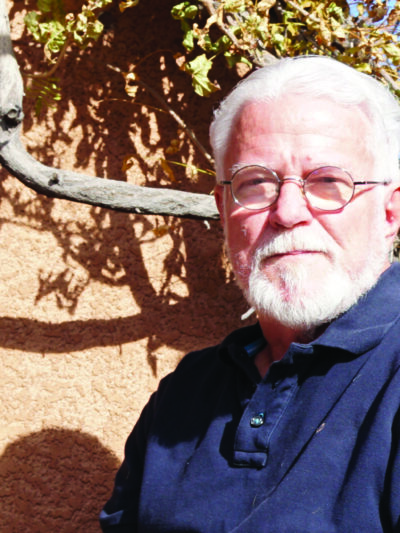 Photo: A man with white hair and beard, wearing glasses sits outside and faces the camera