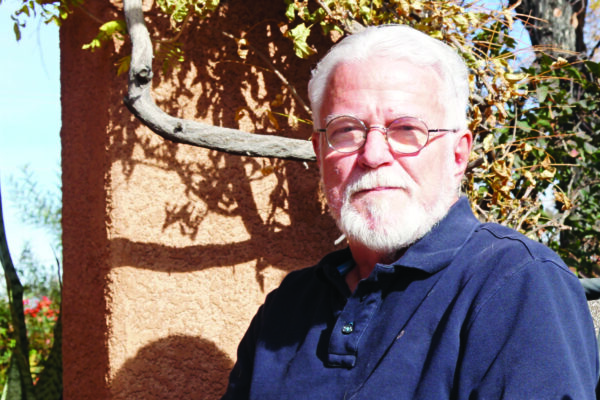 Photo: A man with white hair and beard, wearing glasses sits outside and faces the camera