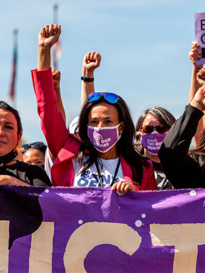 Three women with raised fists, wearing masks with "BAN OFF OUR BODIES" printed on them and carrying a purple banner, lead other protestors at the Women's March Rally For Abortion Justice In Washington, D.C.