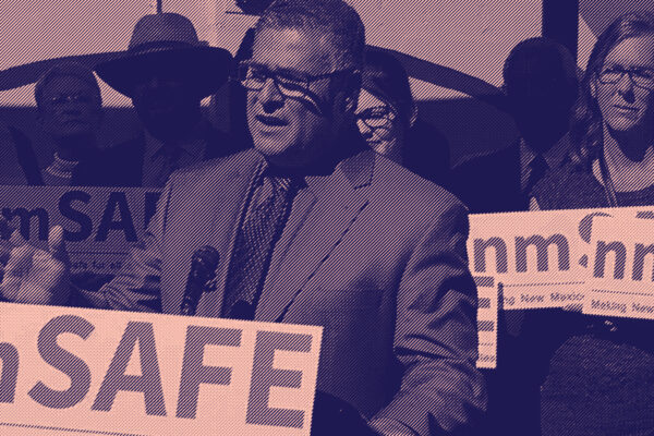 A man speaks at a podium during a public event, surrounded by people holding 'nmSAFE' signs that read 'Making New Mexico safe for all children & families.' The group appears to be advocating for public safety and community well-being in New Mexico