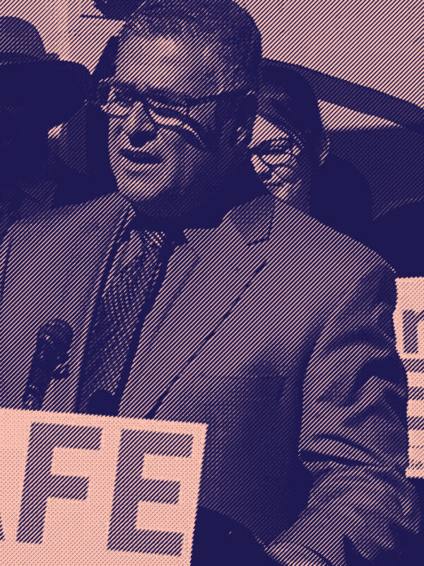 A man speaks at a podium during a public event, surrounded by people holding 'nmSAFE' signs that read 'Making New Mexico safe for all children & families.' The group appears to be advocating for public safety and community well-being in New Mexico