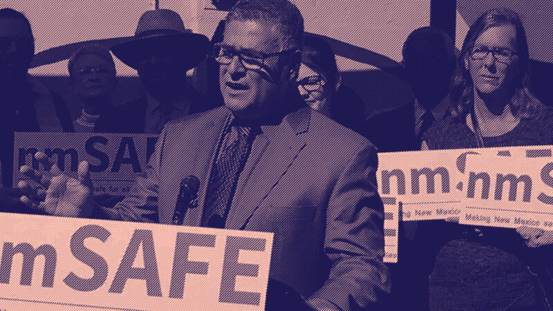A man speaks at a podium during a public event, surrounded by people holding 'nmSAFE' signs that read 'Making New Mexico safe for all children & families.' The group appears to be advocating for public safety and community well-being in New Mexico