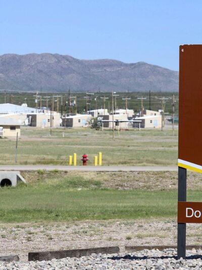 A large brown U.S. Army sign reading “Fort Bliss – Doña Ana Training Complex” stands in the foreground of a wide, open landscape. Behind it are low beige military buildings, utility poles, and a tall red-and-white checkered water tower, with mountains rising in the distance under a clear blue sky.