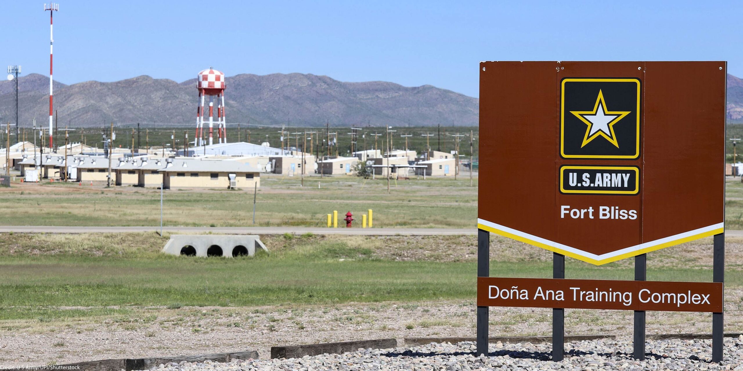 A large brown U.S. Army sign reading “Fort Bliss – Doña Ana Training Complex” stands in the foreground of a wide, open landscape. Behind it are low beige military buildings, utility poles, and a tall red-and-white checkered water tower, with mountains rising in the distance under a clear blue sky.