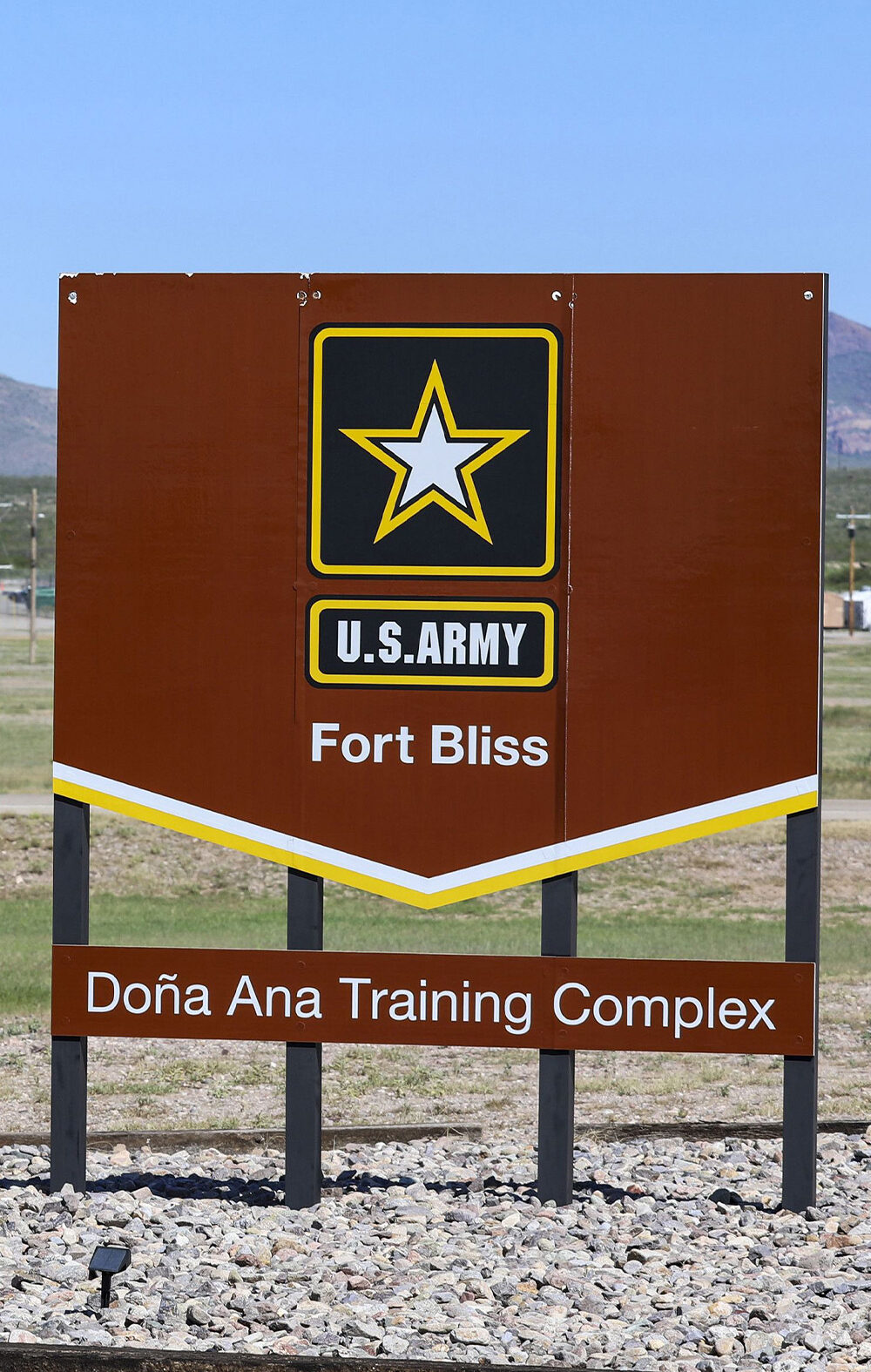 A large brown U.S. Army sign reading “Fort Bliss – Doña Ana Training Complex” stands in the foreground of a wide, open landscape. Behind it are low beige military buildings, utility poles, and a tall red-and-white checkered water tower, with mountains rising in the distance under a clear blue sky.