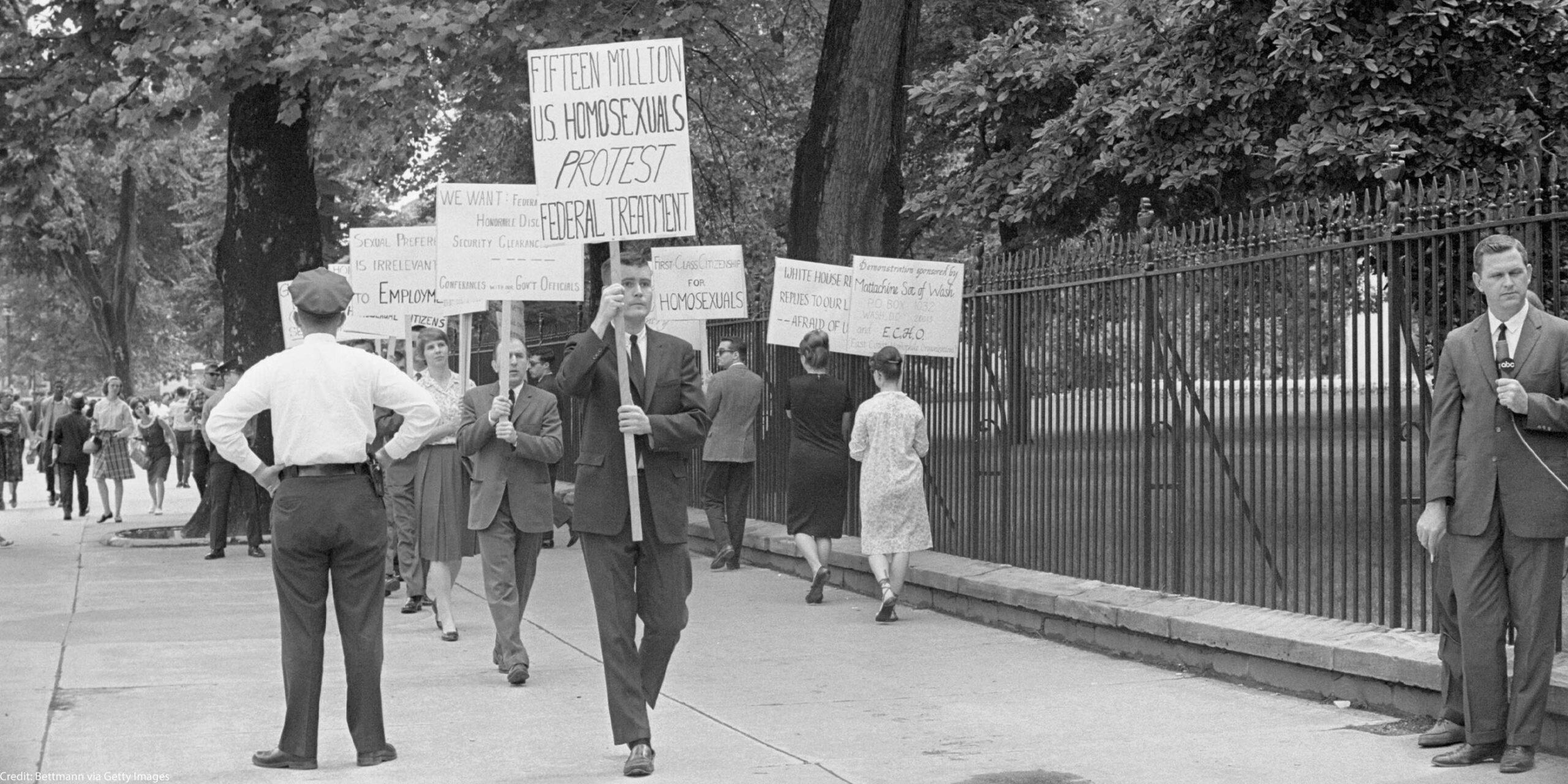 Black-and-white photo of an LGBTQ rights picket on a tree-lined sidewalk beside a tall iron fence (near the White House). Protesters—mostly in suits and dresses—carry hand-lettered signs including “FIFTEEN MILLION U.S. HOMOSEXUALS PROTEST FEDERAL TREATMENT,” “FIRST-CLASS CITIZENSHIP FOR HOMOSEXUALS,” and “SEXUAL PREFERENCE IS IRRELEVANT TO EMPLOYMENT.” A uniformed police officer stands with hands on hips facing the group, while an ABC TV reporter in a suit holds a microphone at the curb; pedestrians walk past in the background.