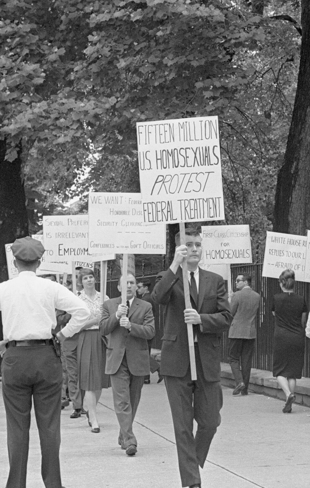 Black-and-white photo of an LGBTQ rights picket on a tree-lined sidewalk beside a tall iron fence (near the White House). Protesters (mostly in suits and dresses) carry hand-lettered signs including “FIFTEEN MILLION U.S. HOMOSEXUALS PROTEST FEDERAL TREATMENT,” “FIRST-CLASS CITIZENSHIP FOR HOMOSEXUALS,” and “SEXUAL PREFERENCE IS IRRELEVANT TO EMPLOYMENT.” A uniformed police officer stands with hands on hips facing the group, while an ABC TV reporter in a suit holds a microphone at the curb; pedestrians walk past in the background.