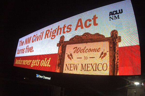 A billboard that says "The NM Civil Rights Act turns five. Justice never gets old" with a "Welcome to New Mexico" sign on it.