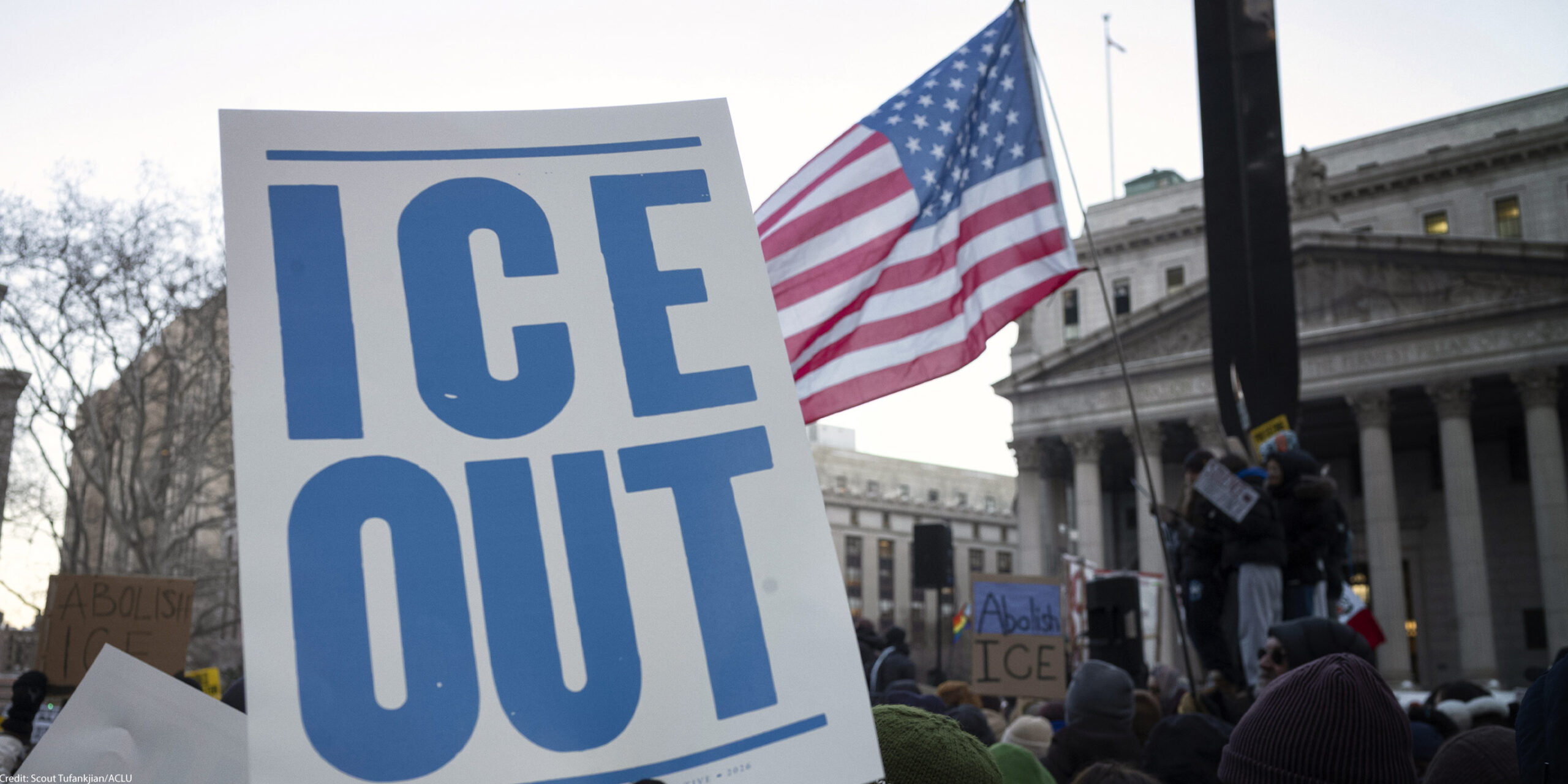 A hand holds a large white protest sign reading "ICE OUT" in bold blue letters during a crowded demonstration outside a government building. An American flag waves above the crowd, and additional signs in the background include messages calling to abolish ICE.