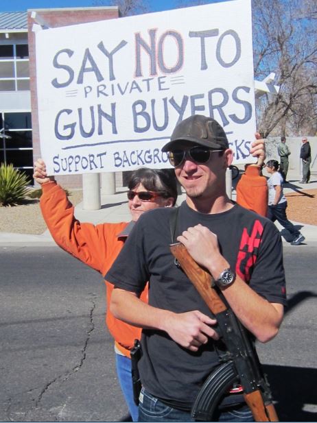 Jill Gatwood holding a homemade protest sign that reads "Say No to Private Gun Buyers. Support Background Checks." She stands behind a man holding a rifle.