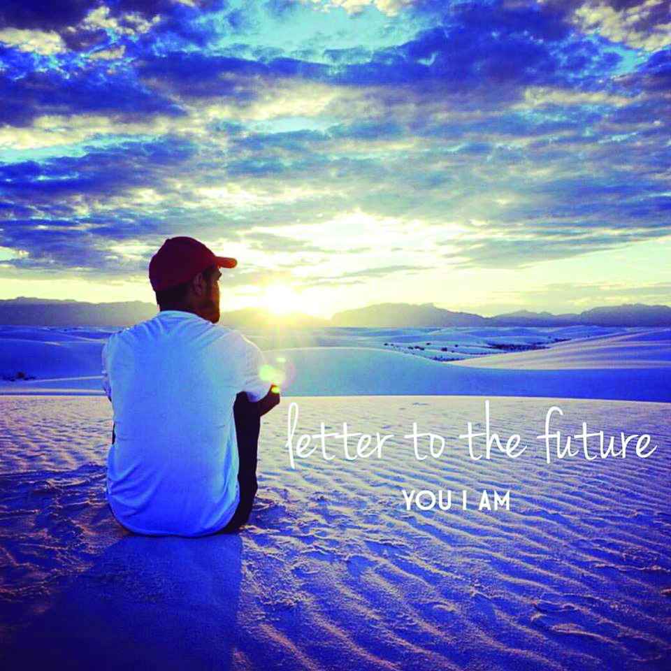 a young man wearing a baseball hat sits on rippled sand with back facing, looking into a distant sunset