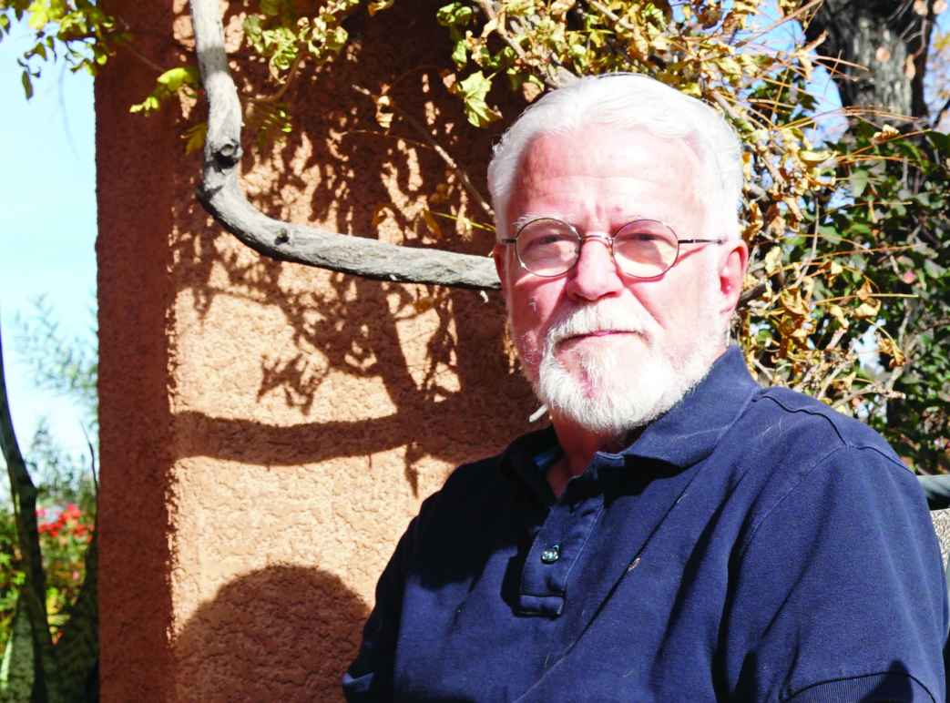 Photo: A man with white hair and beard, wearing glasses sits outside and faces the camera