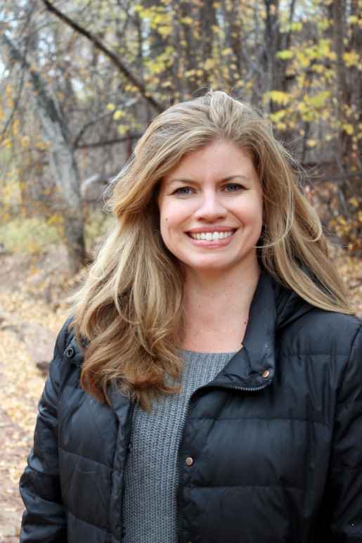 Photo: A woman with blonde hair, smiling, looking at the camera with trees in the background