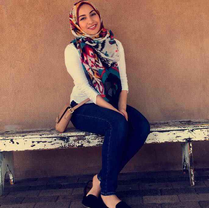 a young women wearing a multi colored hijab, a white long sleeve shirt,and jeans sits on a bench and smiles at the camera