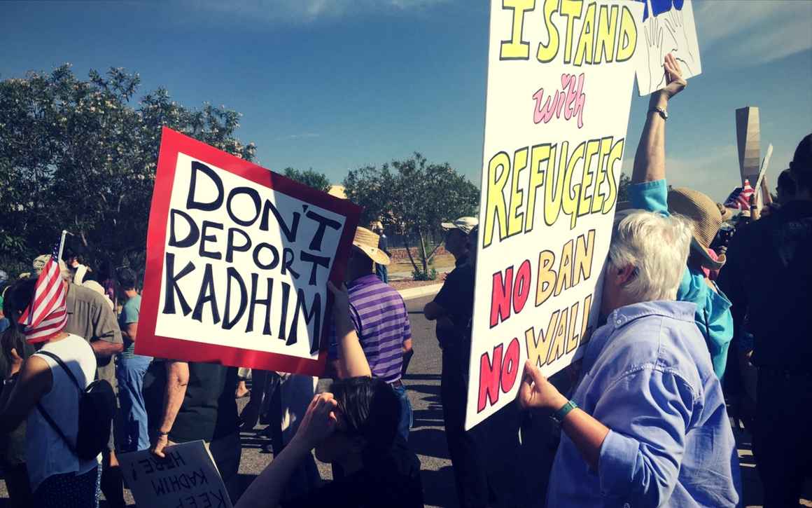 Protesters hold posters that read "Don't deport Kadhim" and "I stand with refugees.  No ban. No wall."