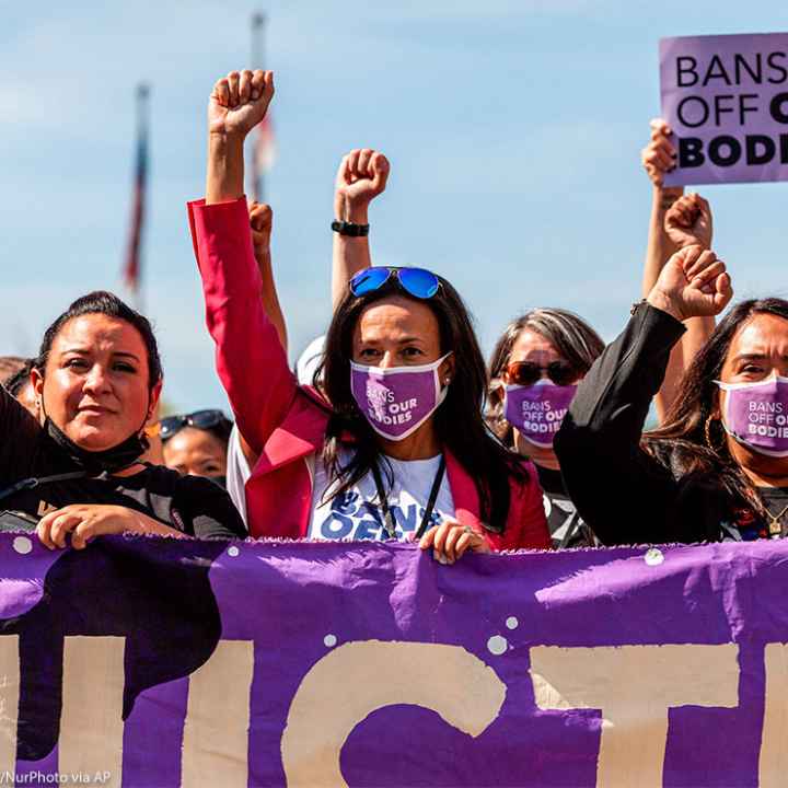 Three women with raised fists, wearing masks with "BAN OFF OUR BODIES" printed on them and carrying a purple banner, lead other protestors at the Women's March Rally For Abortion Justice In Washington, D.C.