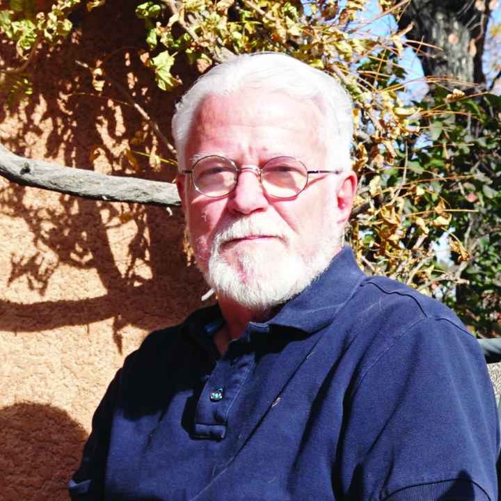Photo: A man with white hair and beard, wearing glasses sits outside and faces the camera