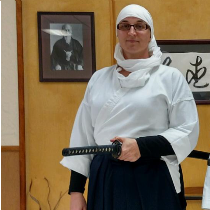 A woman stands wearing a white headscarf and martial arts robe, holding a sword